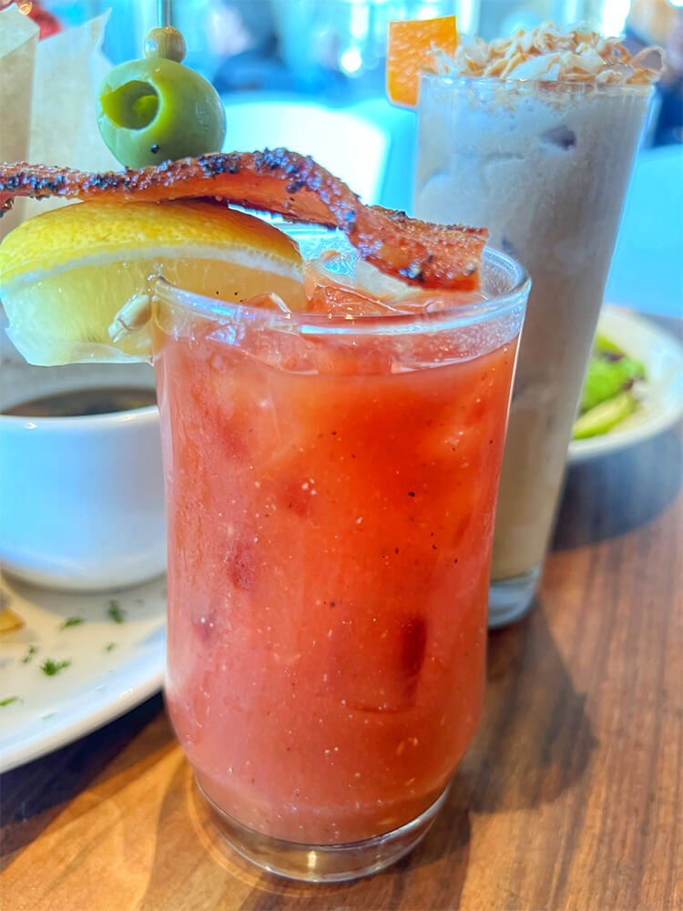 A house-made bloody Mary garnished with a wedge of lemon, peppered bacon, and green olive.