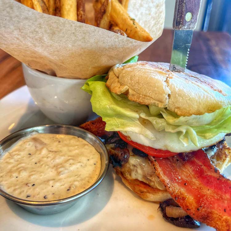 A thick strip of peppered bacon peaks out between lettuce, tomato, grilled onions and melting pepper jack over a grilled sirloin burger on a toasted English muffin. 