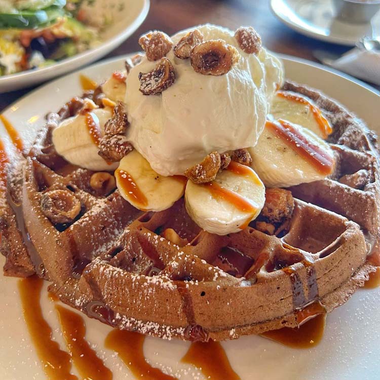 A scoop of vanilla bean ice-cream nestles between slices of banana on a large chocolate waffle topped with a drizzle of caramel and toasted hazelnuts.