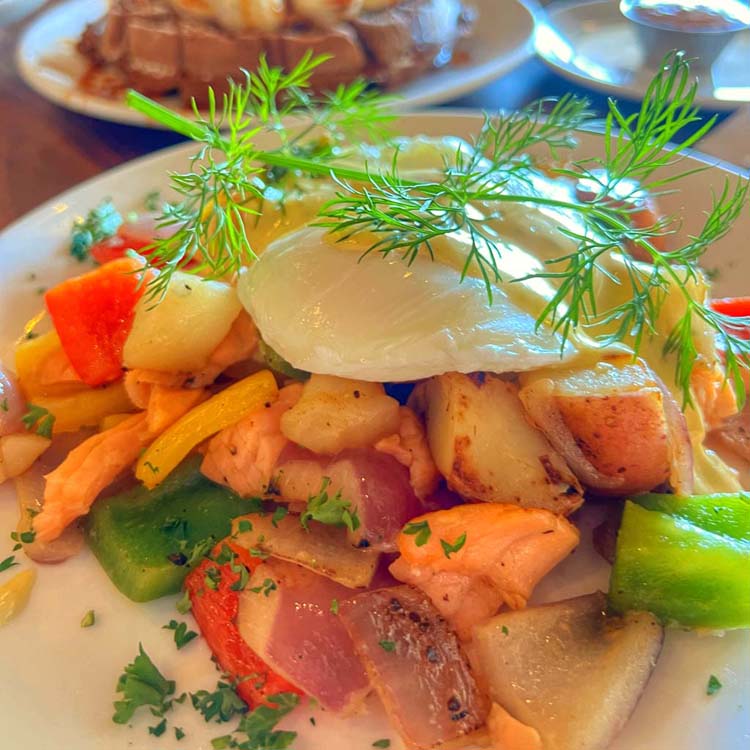 Hunks of smoked salmon and wedges of sautéed onion, bell pepper and red potatoes heap on a plate topped with two poached eggs and dill hollandaise.