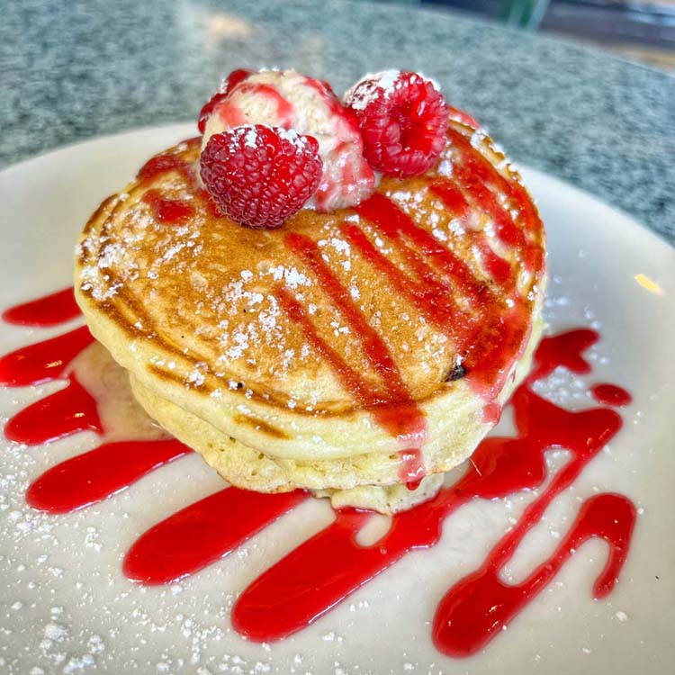 Fresh raspberries and vanilla ice cream sit atop a stack of fluffy pancakes drizzled in raspberry syrup. 