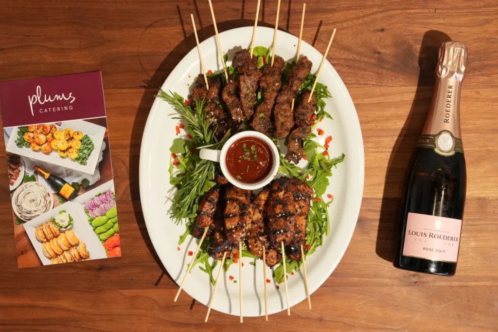 Grilled meat skewers with dipping sauce, catering brochure, and rosé